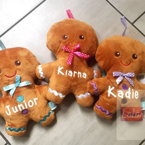 May include: Three plush gingerbread man toys with embroidered details. Each has a name embroidered on the front: "Junior", "Kiarna", and "Kadie". They have colourful bows and ribbons. The toys are brown with white, blue, purple, and pink accents.