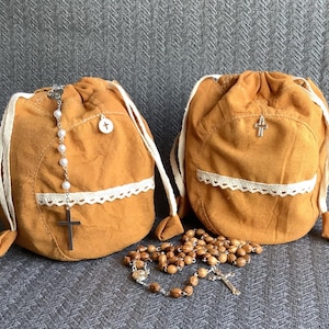 May include: Two mustard-colored drawstring bags with white lace trim and a small pocket. One bag has a pearl rosary and a silver cross, the other has a silver cross. A wooden bead rosary lies in front of the bags.