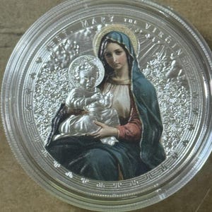 May include: A silver coin featuring a depiction of the Virgin Mary holding the baby Jesus. The coin has a clear, protective casing. The words "ST. MARY THE VIRGIN" are inscribed around the edge of the coin.