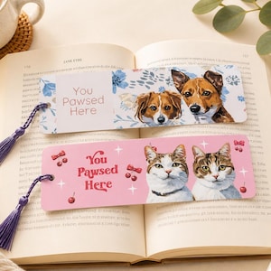 May include: Two decorative bookmarks with tassels rest on an open book. One bookmark features dog illustrations and the text "You Pawsed Here." The other bookmark has cat illustrations and the same text. Both bookmarks have a hole for the tassel.