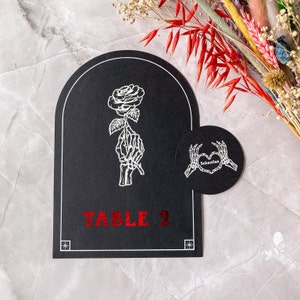 May include: Black table card with a white skeleton hand holding a rose. The card has a rounded top and the text "TABLE 2" in red foil. A smaller, round card features skeleton hands forming a heart with the name "Sebastian".