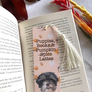 May include: A bookmark featuring a Shih Tzu dog, pumpkins, and the text "Puppies, Books & Pumpkin Spice Lattes." The bookmark has a cream tassel and is placed on an open book.