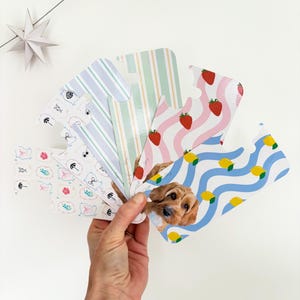 May include: A hand holds a fan of patterned cards in pastel colours. Designs include strawberries, lemons, stripes, and a dog. The cards are on a white background.