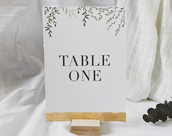 May include: A rectangular table number card with deckled edges, featuring a green watercolor leaf illustration at the top and the black text "TABLE ONE" in the center. The bottom edge is painted with gold leaf, and it rests in a light wood block.