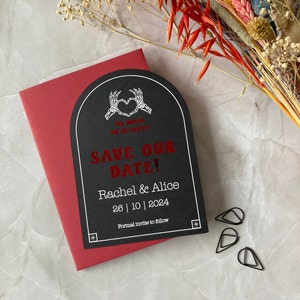 May include: A black and white save the date card with a skeleton hand heart design. The text reads "Til Death Do Us Party!" and "Save Our Date!" with the names Rachel & Alice and the date 26 | 10 | 2024. The card has a red border.