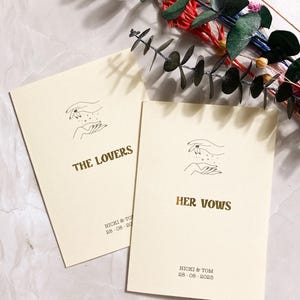 May include: Two cream-coloured cards with gold lettering. The top card says "THE LOVERS" and the bottom card says "HER VOWS". Both cards feature a line drawing of two hands holding a scattering of stars. The date "28.08.2025" is printed on both cards.