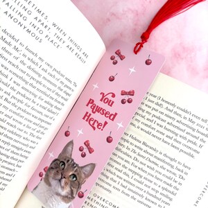 May include: A pink bookmark with a cat illustration and the text "You Pawsed Here!" in red and white lettering. The bookmark is decorated with red cherries and white stars.