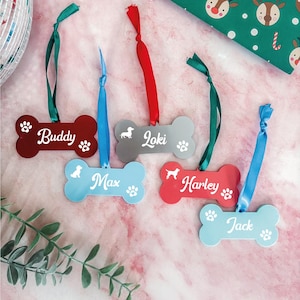 May include: Bone-shaped pet ornaments in red, gray, and blue with names "Buddy", "Loki", "Max", "Harley", and "Jack". Each ornament has a paw print and a ribbon for hanging. A green and white gift wrap with reindeer is in the background.