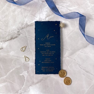 May include: A navy blue menu card with gold foil lettering and stars. The menu includes a starter, main, dessert, and signature cocktails. The text reads: "Menu", "Starter", "Prawn and Crayfish Cocktail with Caviar", "Main", "Traditional Beef Wellington with Roasted Root Veg and a Red Wine Jus", "Dessert", "Lemon Posset with Raspberry Coulis", "Signature Cocktail", "Jessica - Classic Daiquiri", "Liam - Old Fashioned".
