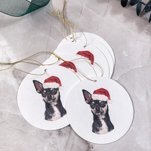 May include: Five round white gift tags featuring a black and white illustration of a chihuahua wearing a red Santa hat. The tags are strung with gold twine.