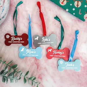 May include: Bone-shaped Christmas ornaments in red, gray, and blue with names and "First Christmas 2025" text. Each ornament has a ribbon for hanging. A green gift wrap with reindeer and a disco ball are in the background.