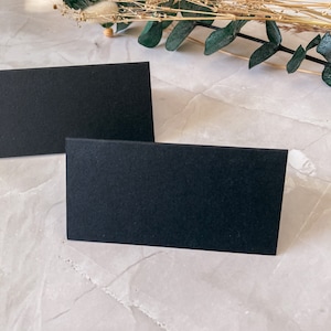 May include: Two black rectangular cards on a white textured surface. The cards are blank and have a matte finish. Dried flowers and greenery are in the background.