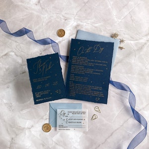 May include: A navy blue wedding invitation suite with gold foil lettering. The invitation card features the text "Thomas & Nicole" and the date "10-06-23". The RSVP card has a gold foil star and a gold foil crescent moon.
