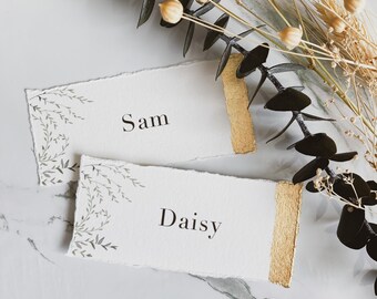 May include: Two rectangular place cards with deckled edges, featuring the names Sam and Daisy printed in black serif font. Each card displays a delicate green botanical illustration on the left and a textured gold painted edge on the right.