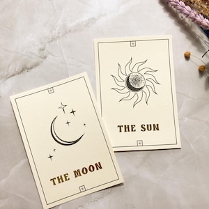 May include: Two tarot cards with cream-colored backgrounds. One card features a crescent moon and stars, labeled "THE MOON." The other card shows a sun with a crescent moon in the center, labeled "THE SUN." Both cards have a black border and gold text.