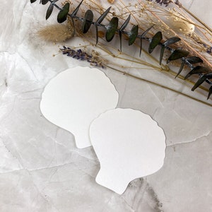 May include: Two white paper seashell-shaped cards on a grey textured background with dried flowers and eucalyptus.
