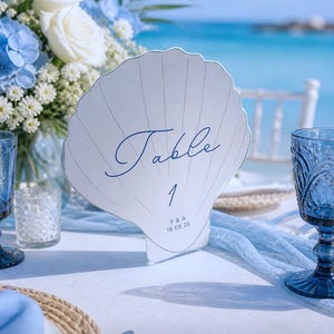 May include: A seashell-shaped table number card with the word "Table" and the number "1" in elegant blue script. The card also includes initials and a date. The card is set on a white tablecloth with blue accents, with a beach and ocean background.