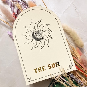 May include: A beige card with a rounded top and a black and white illustration of the sun with a crescent moon inside. The text "THE SUN" is printed in gold foil on the card.