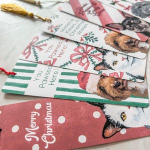 May include: A collection of Christmas-themed bookmarks featuring watercolor illustrations of cats and dogs. The bookmarks have festive designs with text like "Merry Christmas" and "You Pawsed Here!" Some have tassels.