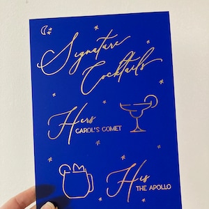 May include: A blue sign with gold lettering that says "Signature Cocktails". The sign has two drink icons with the text "Hers CAROL'S COMET" and "His THE APOLLO".