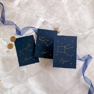 May include: Three navy blue cards with gold foil constellation illustrations and names. The constellations are: Lyra, Pisces, and Monoceros.