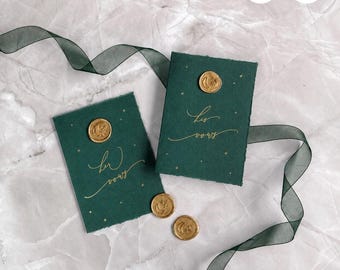 May include: Two rectangular dark green vow booklets with deckled edges, featuring gold foil text reading "her vows" and "his vows." Each booklet is adorned with a gold wax seal and small gold stars, accompanied by a flowing dark green ribbon.