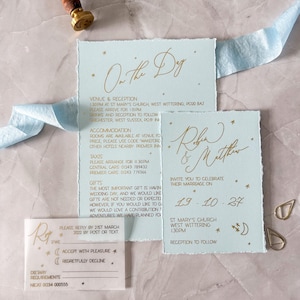 May include: Wedding invitation suite featuring light blue cards with gold calligraphy and star accents. Includes RSVP, details, and invitation cards. A wax seal and blue ribbon add decorative elements. A beautiful set for a wedding.