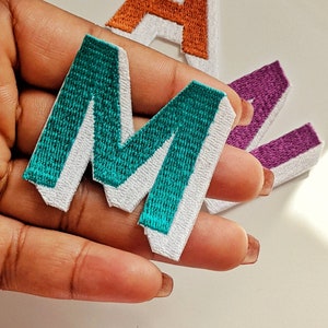 May include: A teal felt letter M with a white outline. The letter is being held in a hand.