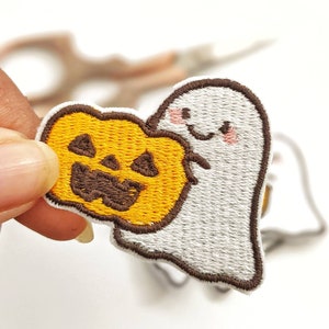 May include: A white ghost with a friendly face is holding a yellow and orange pumpkin with a carved jack-o'-lantern face. The embroidered patch is perfect for adding a touch of Halloween fun to clothing, bags, or crafts.