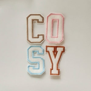 May include: Four felt letters spelling out the word "COSY" in brown, pink, light blue, and brown. The letters are in a varsity style font.
