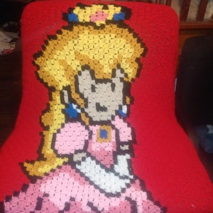May include: A red crocheted chair cover featuring a pixelated image of Princess Peach from the Super Mario video game series. The princess is depicted with blonde hair, a pink dress, and blue earrings.