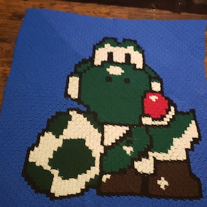 May include: A crocheted blanket featuring a green Yoshi character holding a red apple. The Yoshi is wearing a brown hat and boots. The blanket is made with blue, green, white, red, and brown yarn.