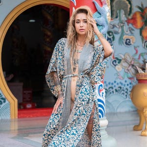 May include: A blue and gold patterned kimono with a long, flowing silhouette. The kimono has a sash tied at the waist and is open in the front, revealing a nude undergarment. The kimono is worn by a person with long, wavy hair and a gold necklace.