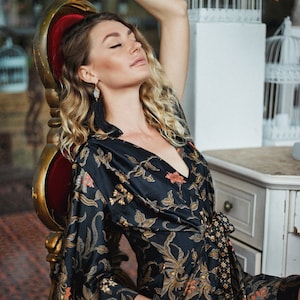 Op de afbeelding: Een vrouw draagt een zwarte en gouden robe met een bloemenprint en een kwast oorring. Ze zit op een rode fluwelen stoel met haar ogen dicht.