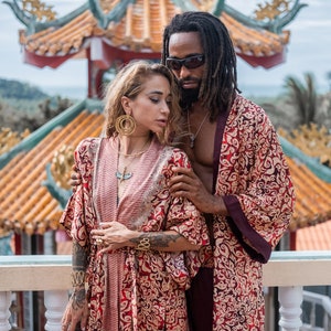 May include: A couple wearing matching red and gold patterned robes stand in front of a traditional Chinese temple. The woman is wearing gold jewelry and the man has dreadlocks and is wearing sunglasses.