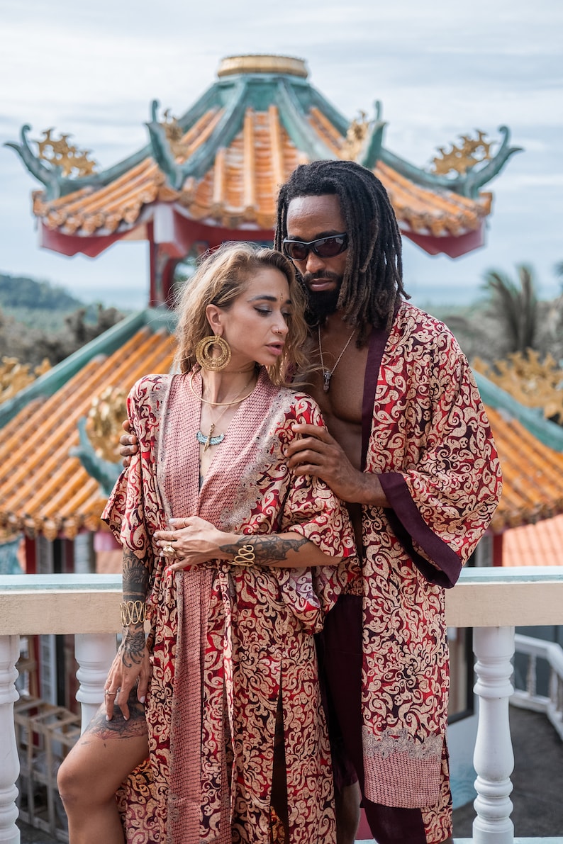May include: A couple wearing matching red and gold patterned robes stand in front of a traditional Chinese temple. The woman is wearing gold jewelry and the man has dreadlocks and is wearing sunglasses.