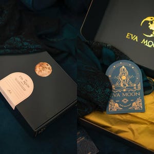 May include: Black gift box with gold foil accents, featuring the "EVA MOON" logo and a moon graphic. The box is open, revealing a gold tissue paper lining and a teal card with a gold illustration. A round sticker with a moon graphic is on the box.