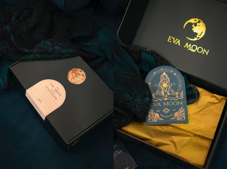 May include: Black gift box with gold foil accents, featuring the "EVA MOON" logo and a moon graphic. The box is open, revealing a gold tissue paper lining and a teal card with a gold illustration. A round sticker with a moon graphic is on the box.