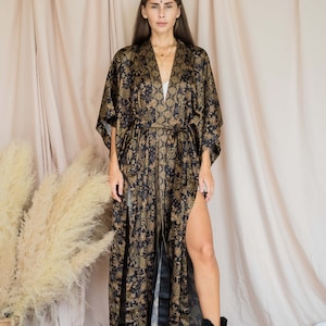 May include: A long, black and gold patterned kimono-style robe with a thigh-high split. The robe features a floral design and is tied at the waist. The model is wearing black boots. The background is a neutral beige.