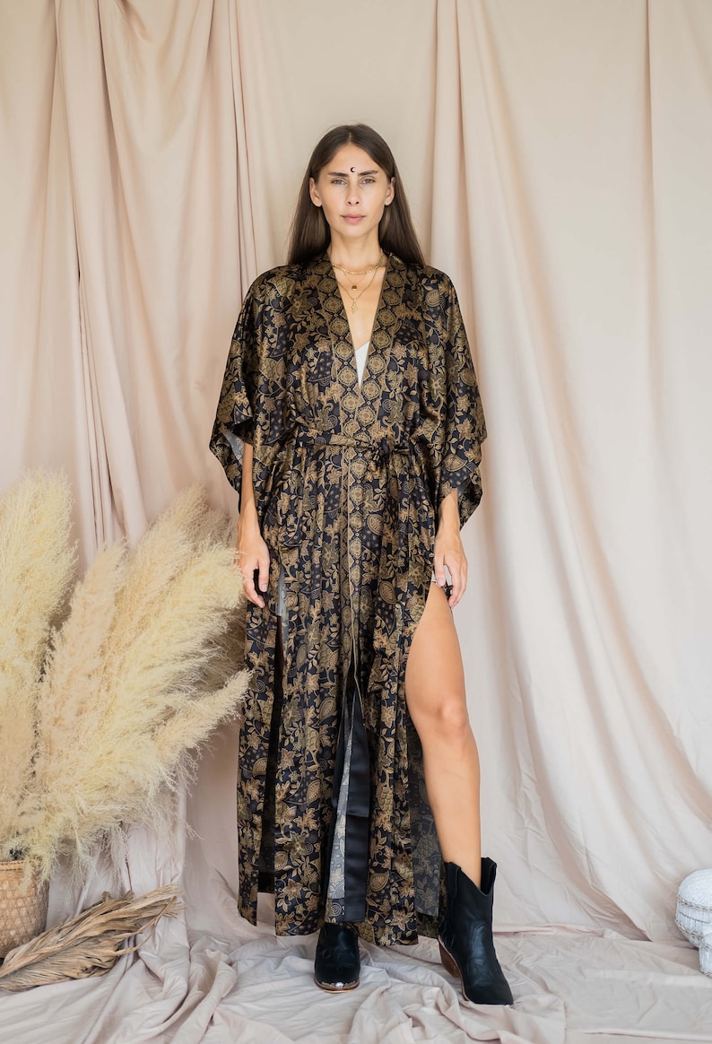May include: A long, black and gold patterned kimono-style robe with a thigh-high split. The robe features a floral design and is tied at the waist. The model is wearing black boots. The background is a neutral beige.