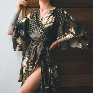 May include: A black and gold patterned wrap robe with wide sleeves. The robe features a batik-style design with floral and geometric motifs. The fabric is dark with gold and white accents. The robe is tied at the waist and has a thigh-high slit.