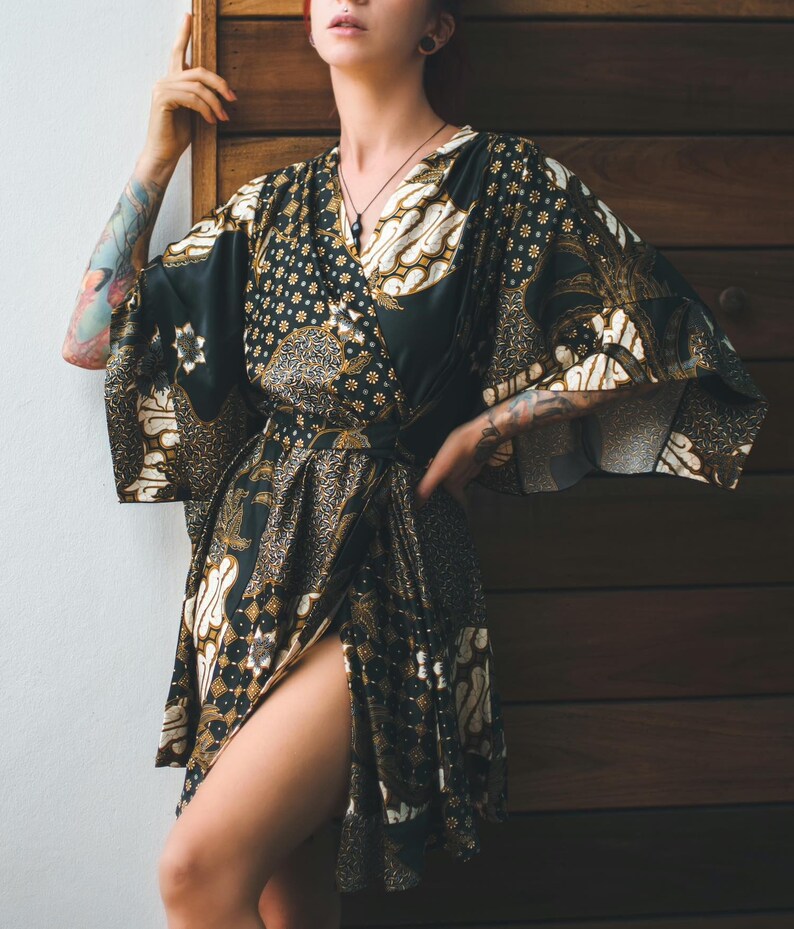 May include: A black and gold patterned wrap robe with wide sleeves. The robe features a batik-style design with floral and geometric motifs. The fabric is dark with gold and white accents. The robe is tied at the waist and has a thigh-high slit.