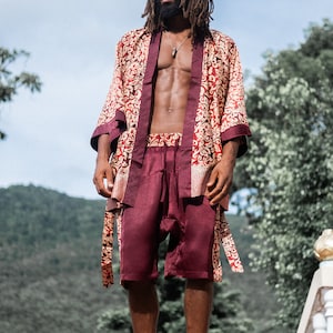 May include: A man wearing a red and gold patterned silk robe with a matching pair of shorts. He is standing in front of a green hillside and wearing sunglasses.