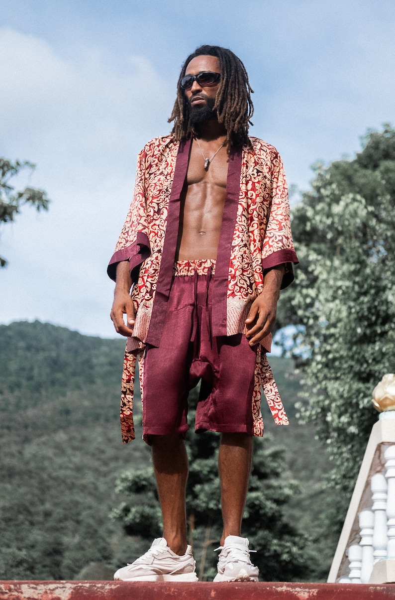 May include: A man wearing a red and gold patterned silk robe with a matching pair of shorts. He is standing in front of a green hillside and wearing sunglasses.