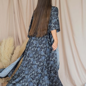 May include: A long, flowing black and blue floral print kimono with a long sleeve. The kimono is being worn by a person with long brown hair. The person is standing in front of a light beige background.