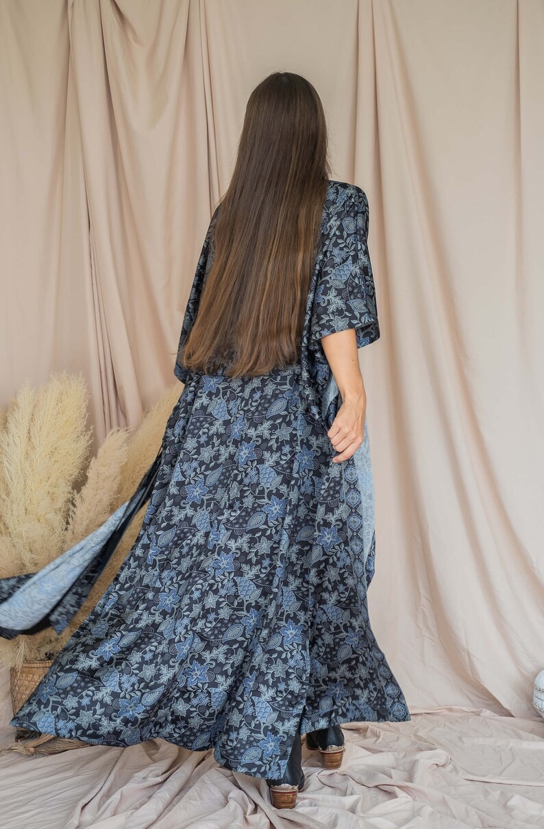 May include: A long, flowing black and blue floral print kimono with a long sleeve. The kimono is being worn by a person with long brown hair. The person is standing in front of a light beige background.