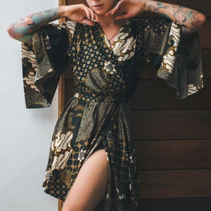 May include: A black and gold patterned wrap dress with a high slit. The dress features a floral and batik-style design. The sleeves are wide and flowing. The dress is tied at the waist.