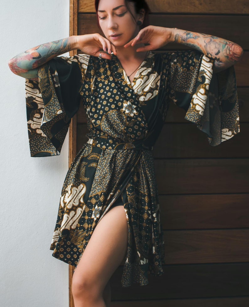 May include: A black and gold patterned wrap dress with a high slit. The dress features a floral and batik-style design. The sleeves are wide and flowing. The dress is tied at the waist.