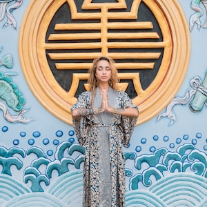 May include: A woman wearing a long, blue and gold patterned robe stands in front of a blue and gold wall with a large circular design. She has her eyes closed and her hands are clasped in front of her.