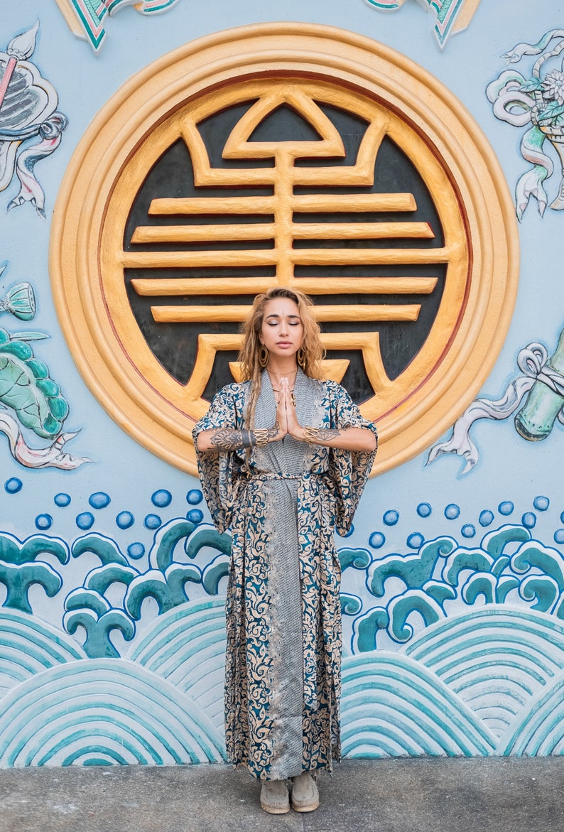 May include: A woman wearing a long, blue and gold patterned robe stands in front of a blue and gold wall with a large circular design. She has her eyes closed and her hands are clasped in front of her.